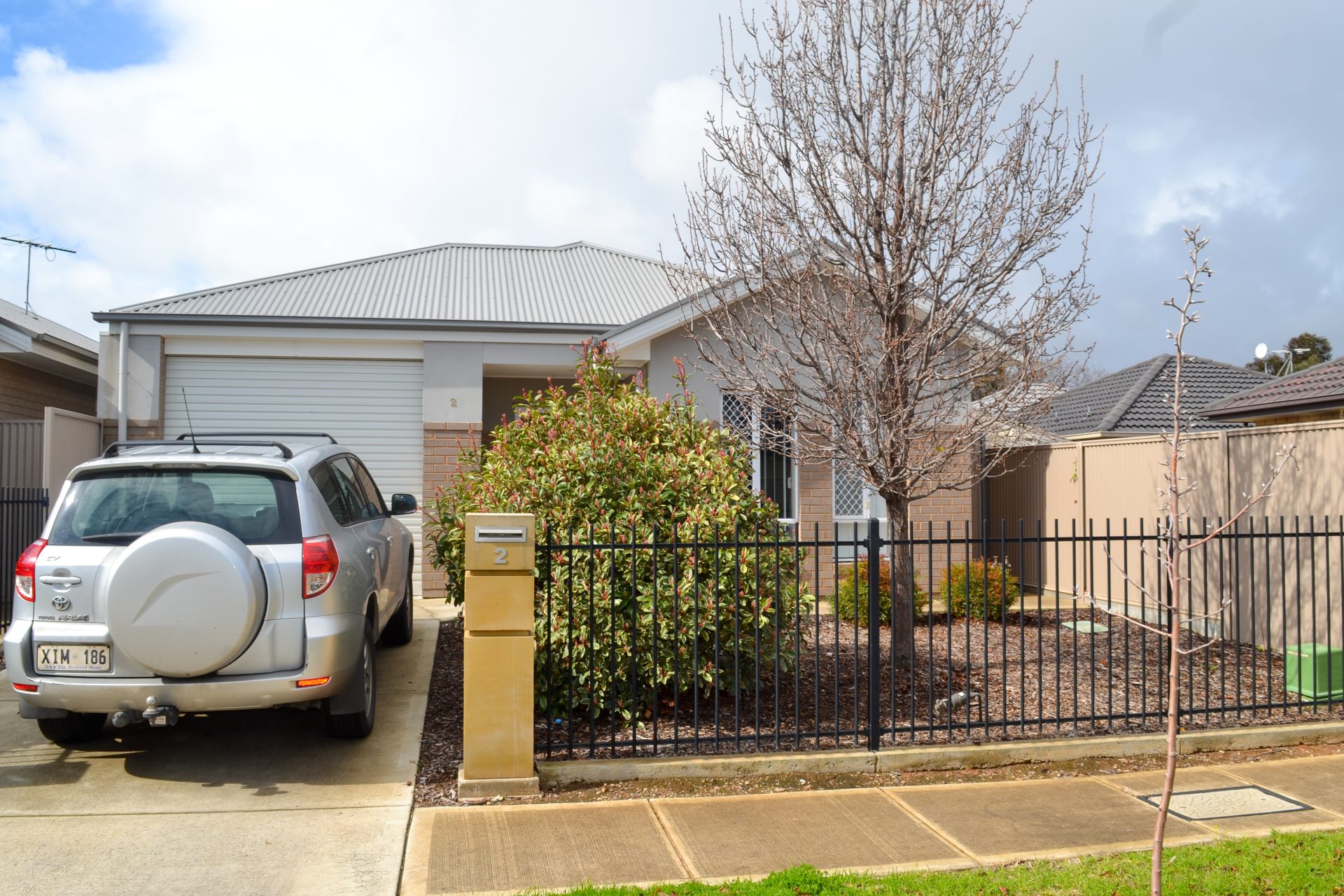Facade of home - beige brick home with grey tin rood. A black post fence with small front yard. 