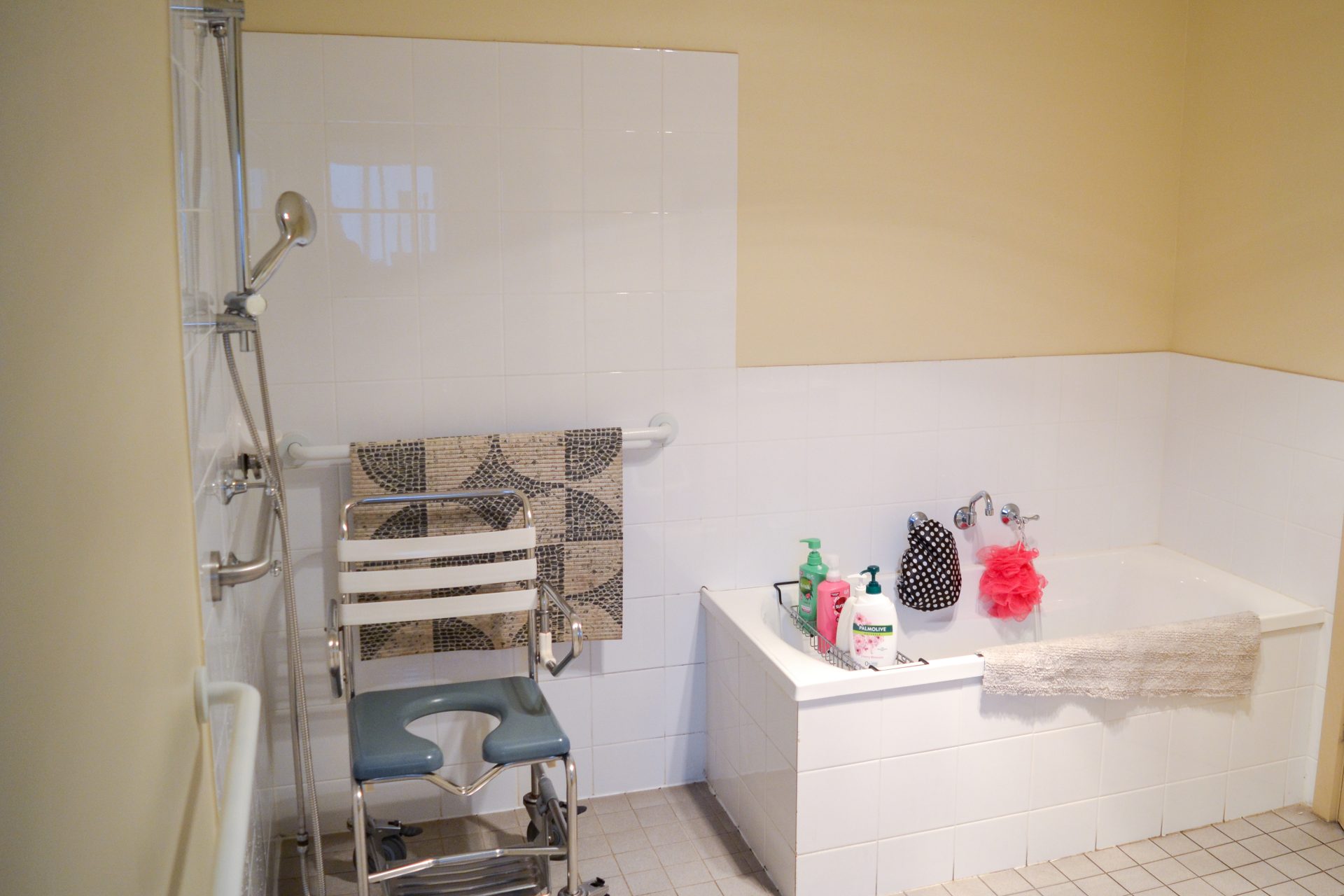 Bbathroom with yellow painted walls and white wall tiles in wet areas. 
Beige floor tiles with safety and shower rails scattered throughout. 