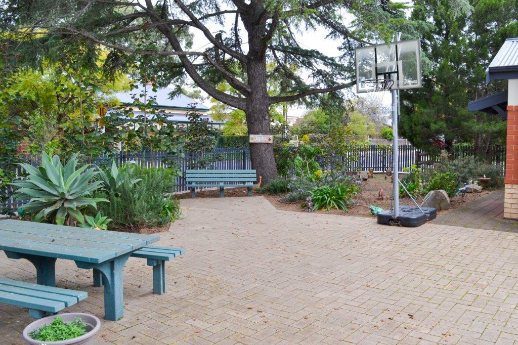 Front garden and entertainment area of Amaroo. Large paved space with picnic tables, plants and basketball ring. A blue picket fence lines the property.