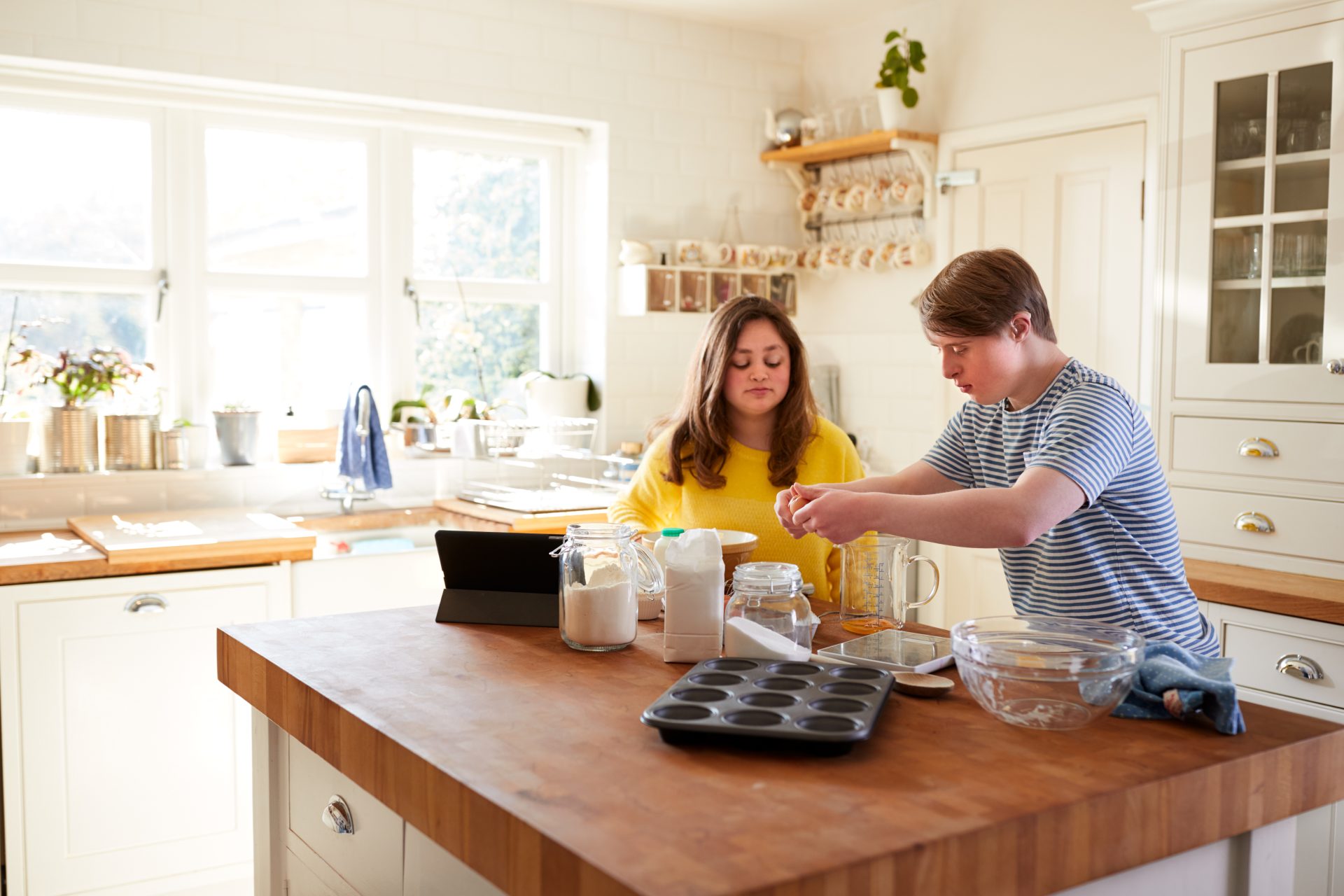 Two people baking muffins in a light and bright kitchen. Male is cracking egg, wearing a blue and white stripe shirt. Female is wearing a yellow knit jumper. 